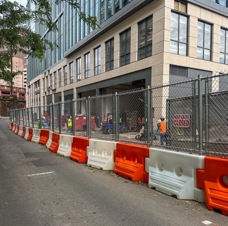 Alternating orange and white WATERCADE barriers with chain-link fence panels along a city street
