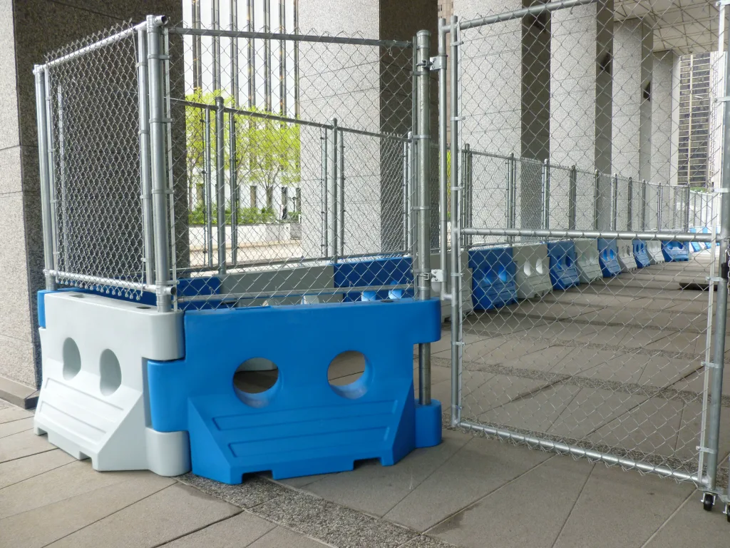 Blue and white WATERCADE barriers with chain-link fence panels around a plaza fountain